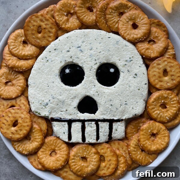 A spooky food dish featuring a skull-shaped cheese ball with black olive eyes and mouth, surrounded by round crackers arranged in a circle.