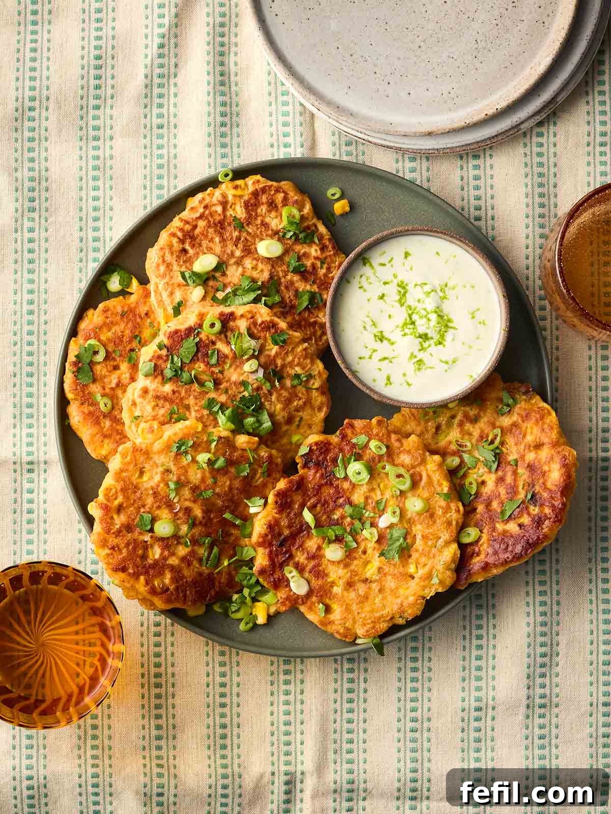 Crispy Sweetcorn Fritters Recipe 2 A plate of cooked sweetcorn fritters with a yoghurt tip and garnished with coriander and spring onions, on a green and white stripy tablecloth.