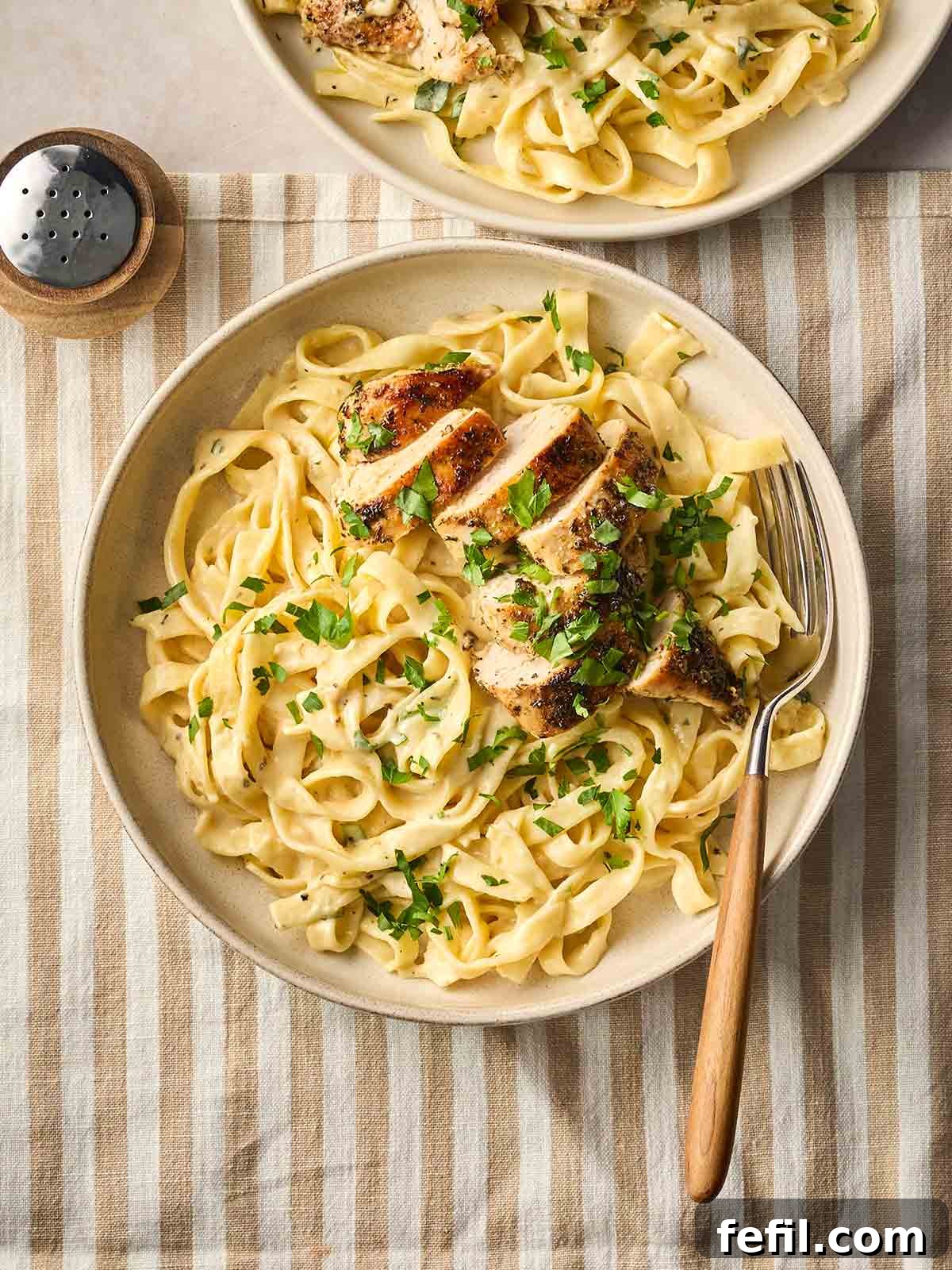 Classic Creamy Chicken Alfredo Recipe 3 A bowl of Chicken Alfredo on a table with a fork, with another bowl to the top of the image, with a stripy placement underneath and pepper to the side.