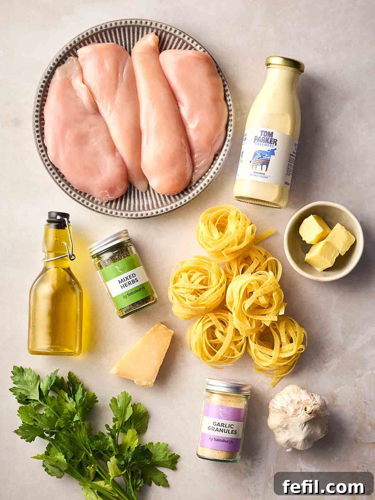 Classic Creamy Chicken Alfredo Recipe 4 Raw ingredients for making Chicken Alfredo laid out on a white counter, including chicken breasts, tagliatelle, cream, oil, butter, parmesan and herbs.