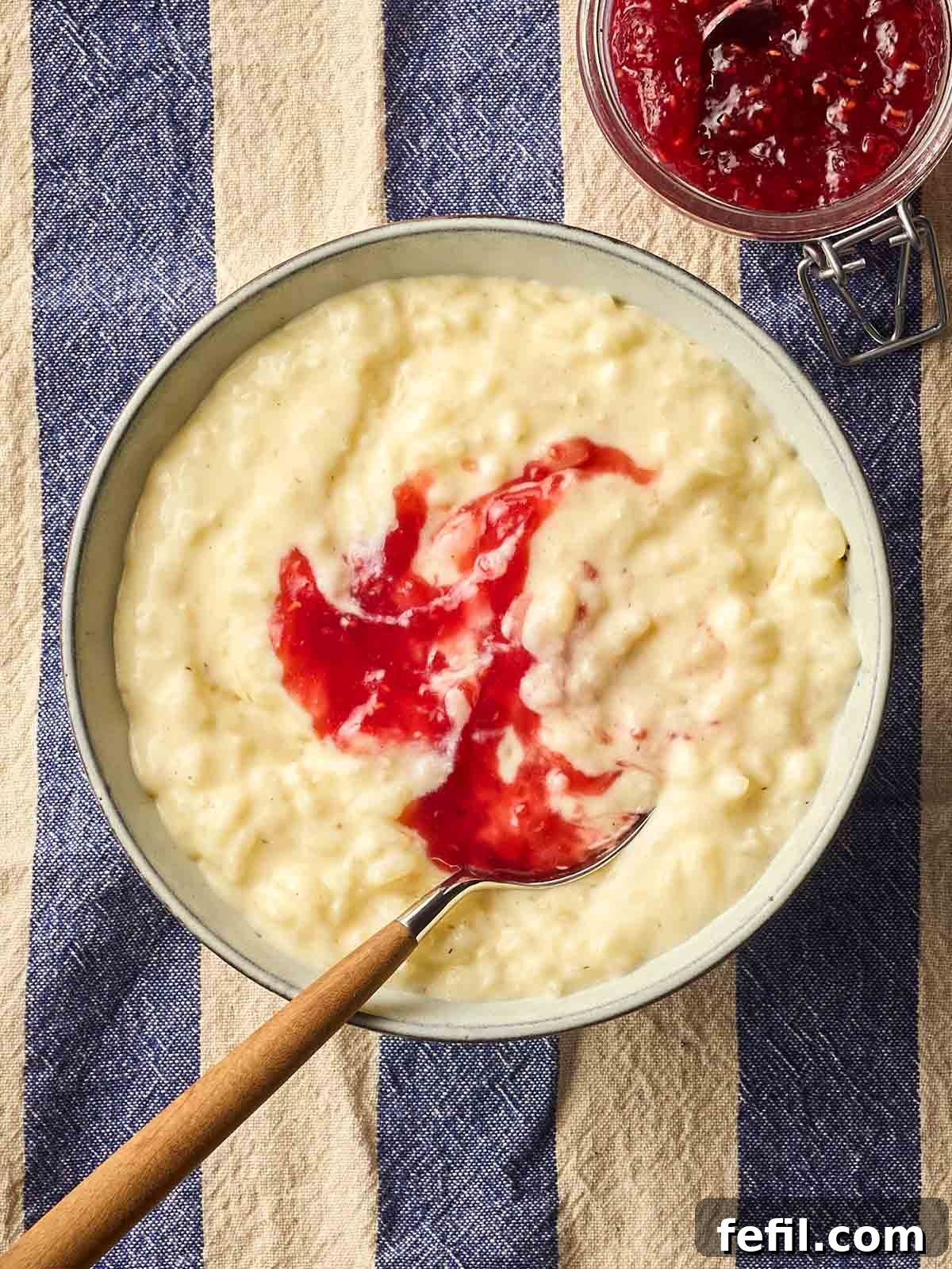 Creamy Slow Cooker Rice Pudding Recipe 3 A bowl of homemade slow cooker rice pudding, with jam on top with a spoon, on a white and blue stripy tablecloth.