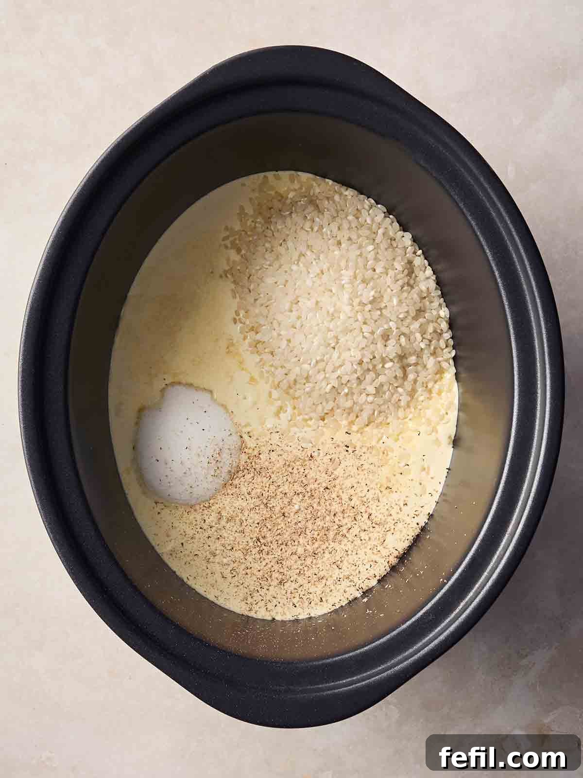 Creamy Slow Cooker Rice Pudding Recipe 5 A slow cooker pan on a white surface with uncooked ingredients for rice pudding inside.