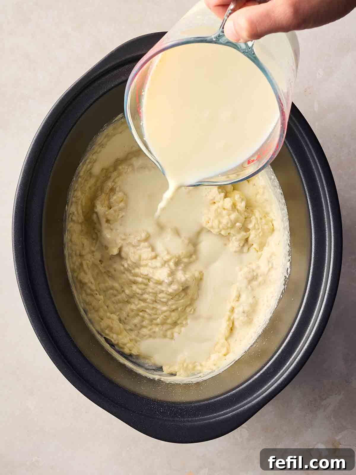 Creamy Slow Cooker Rice Pudding Recipe 6 A crock pot pan with cooked rice pudding inside, with a hand pouring more cream into it.