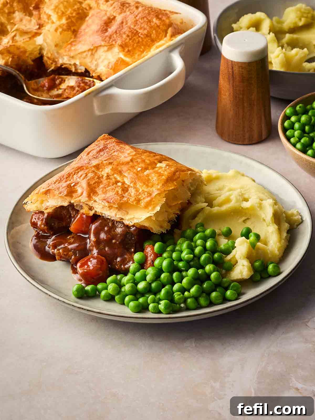 A plate of homemade Steak and Ale Pie with mashed potatoes and peas on a plate, on a dinner table.