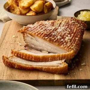 How to Make Crispy Pork Belly with Perfect Crackling Skin 9 A cooked joint of pork belly with crackling on a wooden serving board, with a small pot of apple sauce and a bowl of roast potatoes.